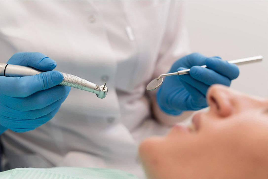 Manos con guantes sostienen la turbina y el espejo dental junto a la boca del paciente