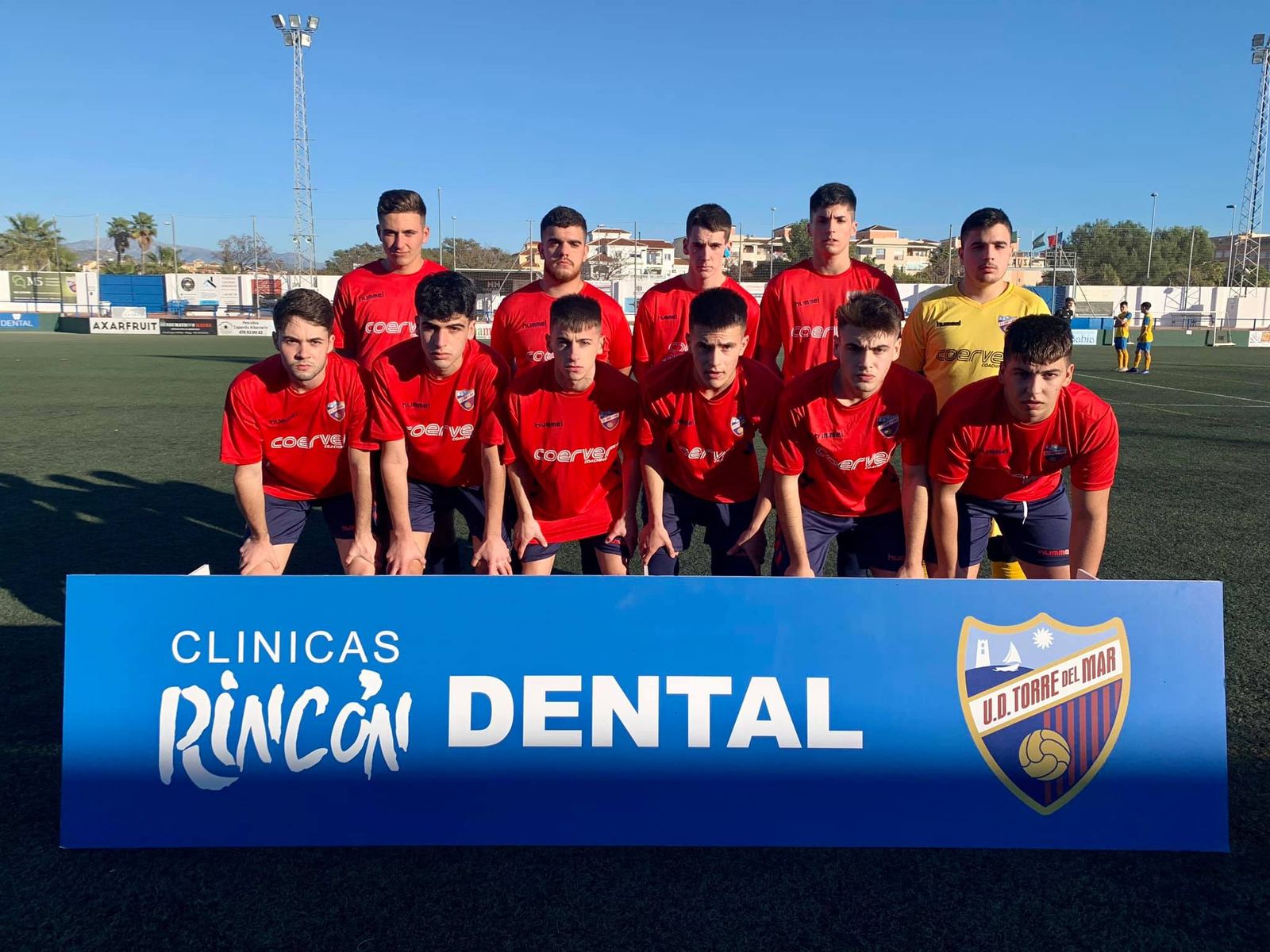 Equipo juvenil de fútbol de la U.D. Torre del Mar posa tras la valla de Clínicas Rincón Dental