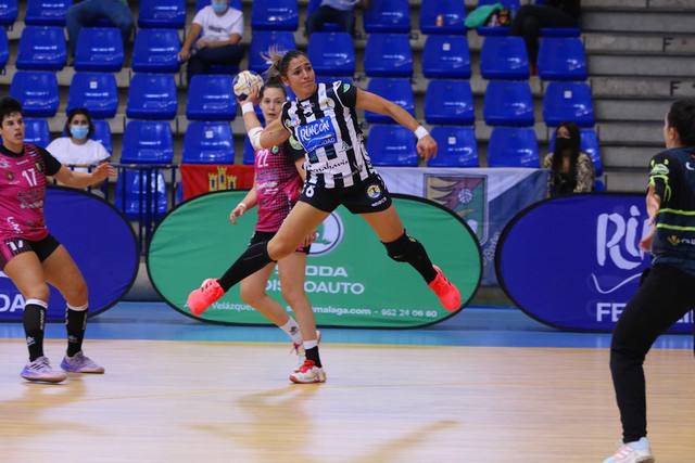 Una jugadora de balonmano salta para lanzar ante la defensa, con la publicidad de Rincón Fertilidad al fondo