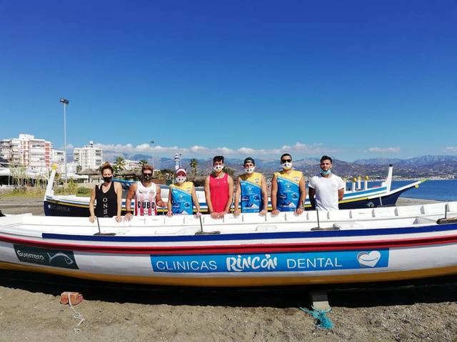 La tripulación posa junto a la jábega en la playa, con el rótulo de Clínicas Rincón Dental en el casco